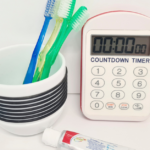 Toothbrushes in cup next to toothpaste and timer.