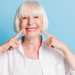 Photo portrait of older woman with blonde hair pointing at white teeth healthy smile dental whitening veneers isolated on bright blue color background.