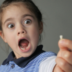 Young girl (female age 6) surprised to find that she have lost her first bottom front milk teeth isolated on grey background.