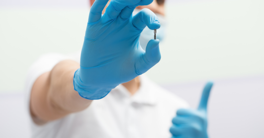 Dentist in a white uniform with blue latex gloves holding a dental implant.