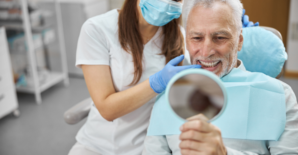 Elderly man and his dentist smiling while looking in the mirror at a dental implant clinic.