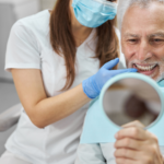 Elderly man and his dentist smiling while looking in the mirror at a dental implant clinic.